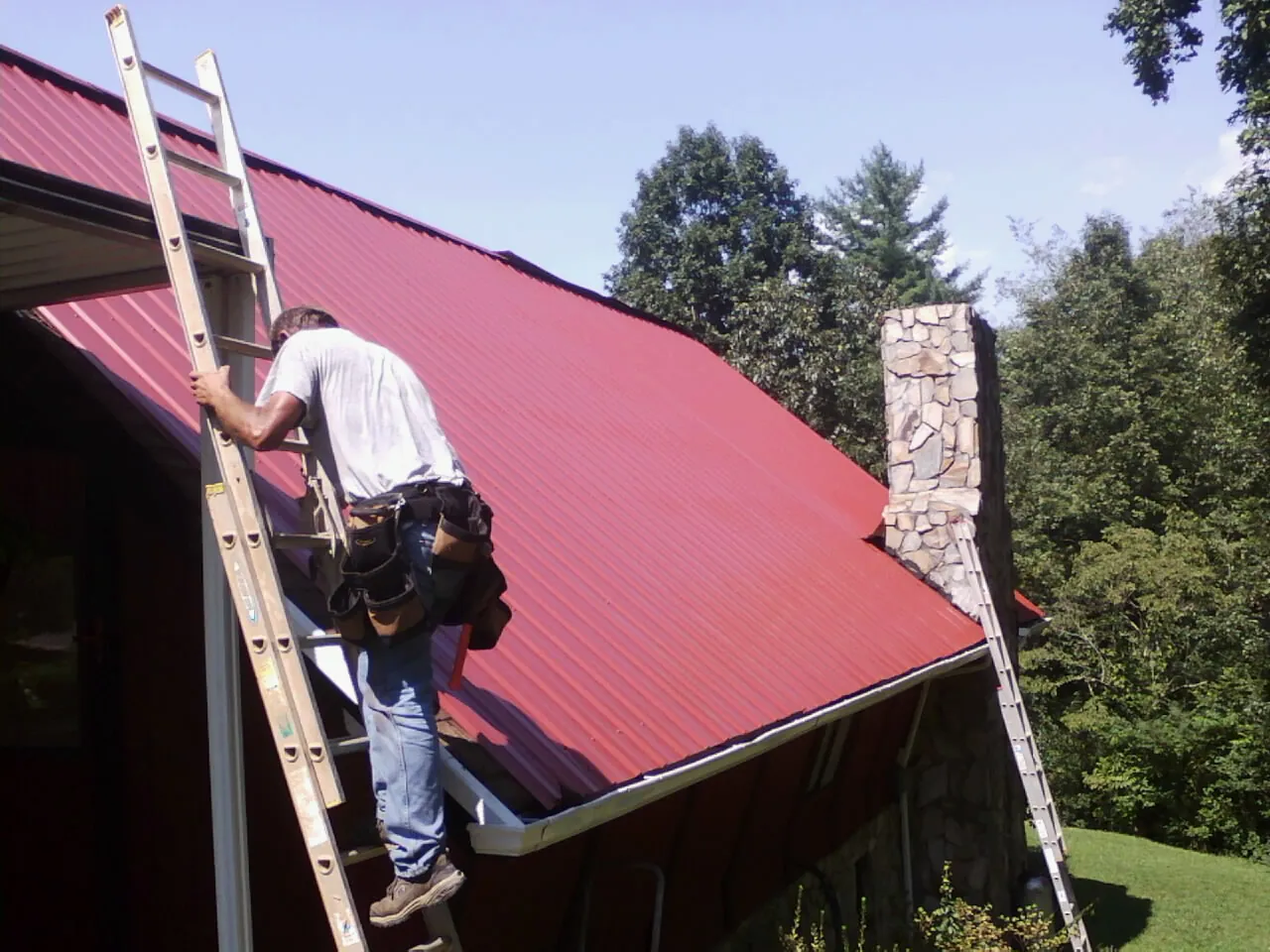 Roofer on a ladder beside a red metal roof and stone chimney — Soffit & Fascia Repair by Roofing Brothers in Whitewater